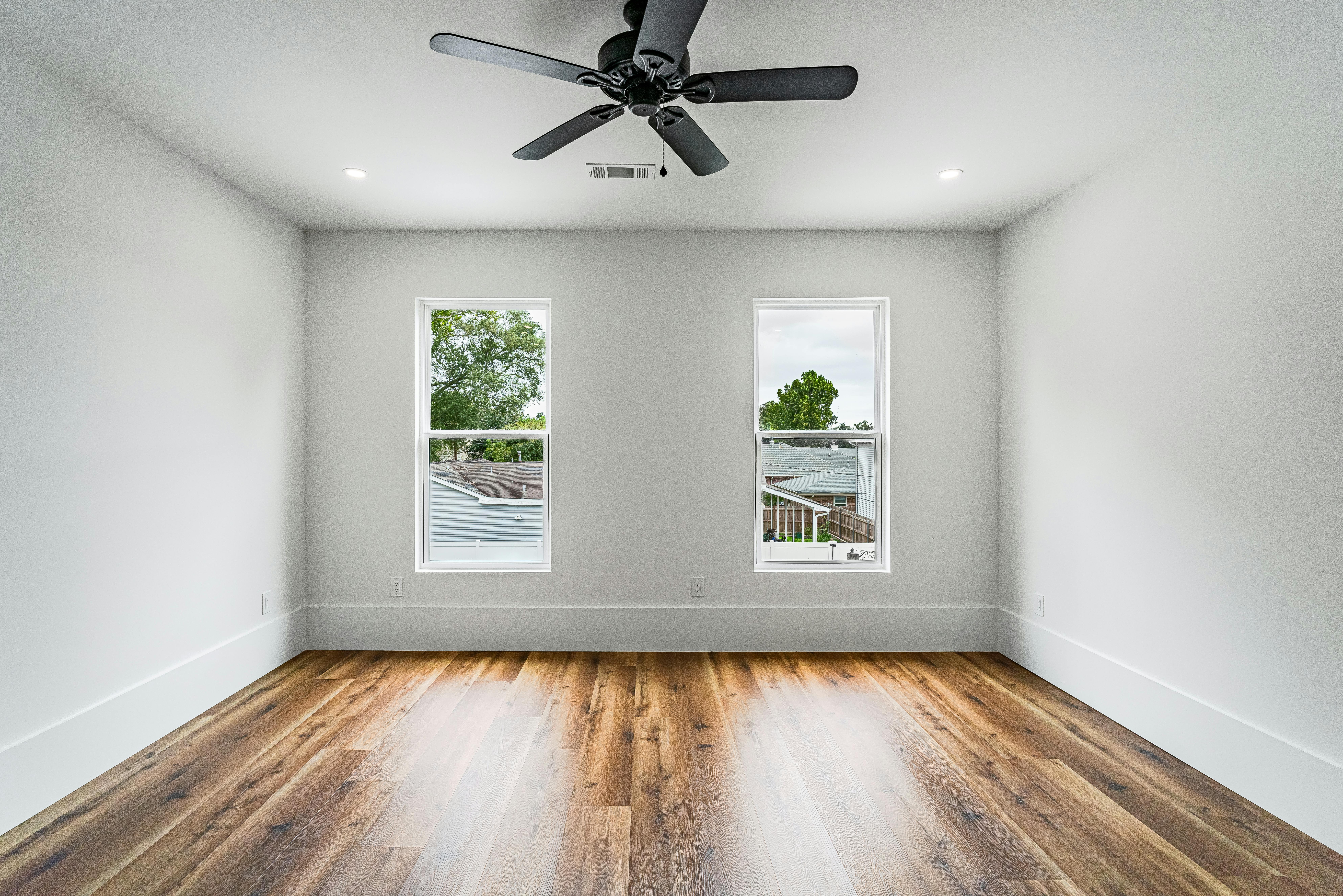 Empty room before staging - gray walls, beautiful hardwood floors, ceiling fan, and two windows with natural light